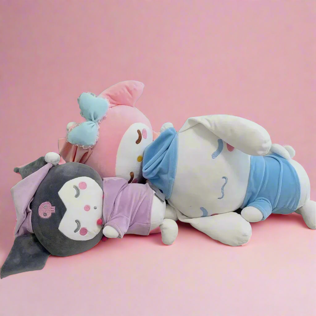 A set of three Sanrio character plushies, including a purple one, a blue one, and a gray one, lying on a bed.