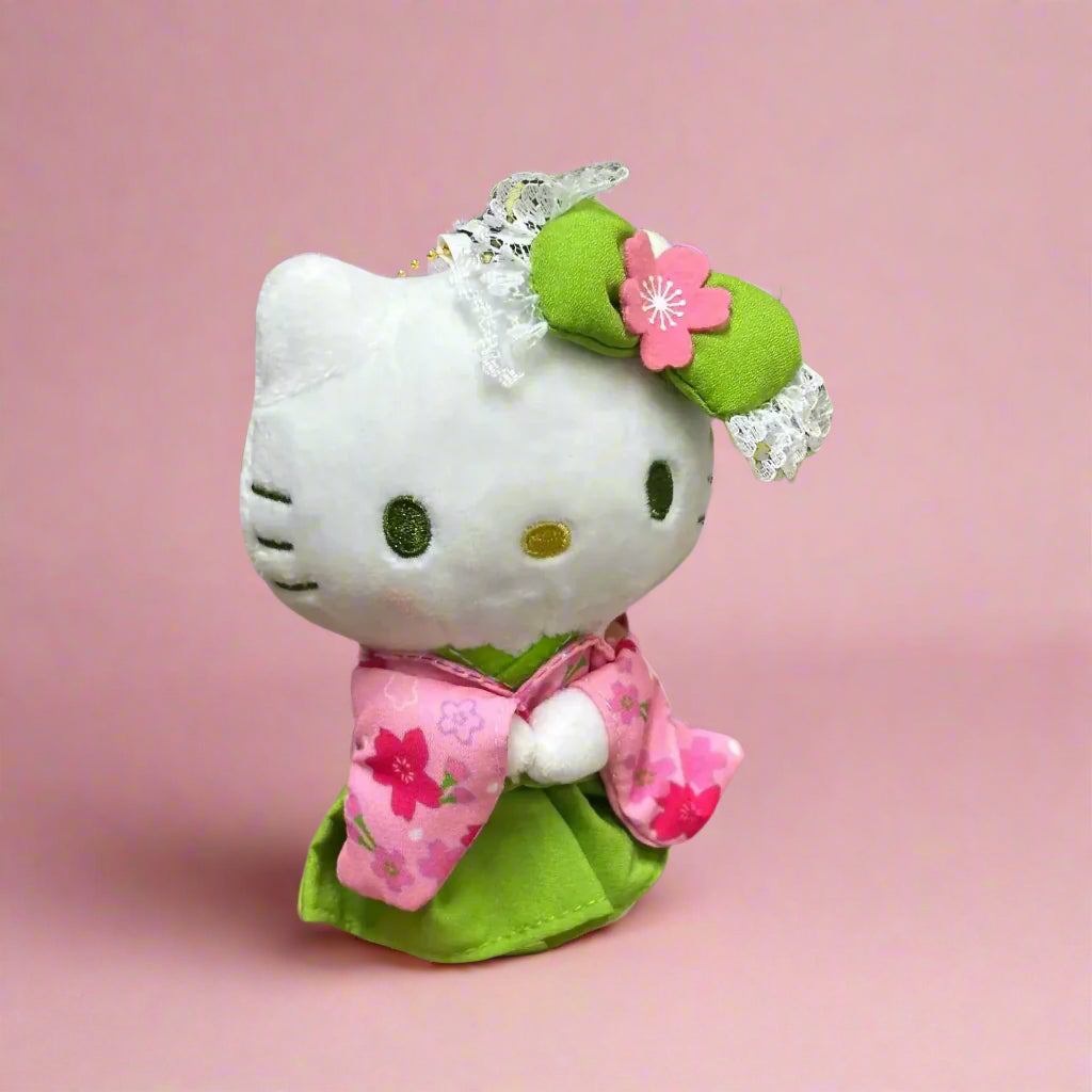 A plushie keychain of Hello Kitty wearing a green kimono with matcha theme, held in front of a shelf with various colorful toy figurines.