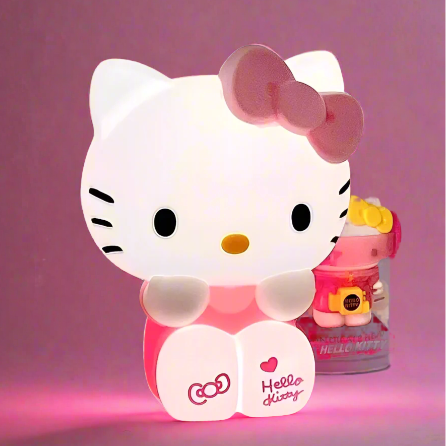 A Hello Kitty themed bedroom night light lamp with the character's face illuminated in pink color. The lamp is accompanied by small Hello Kitty toys and items in the background.