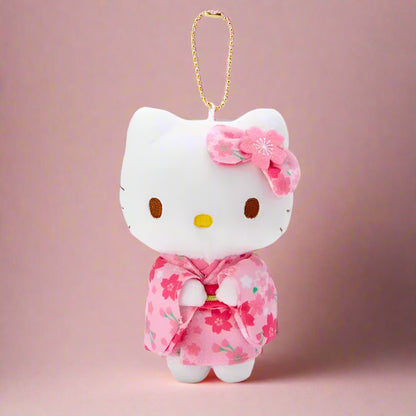 A plushie keychain of Hello Kitty wearing a green kimono with matcha theme, held in front of a shelf with various colorful toy figurines.