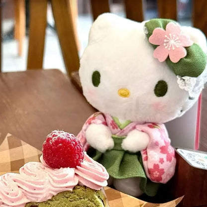 A plush toy of Hello Kitty dressed in a matcha kimono, sitting next to a piece of matcha dessert with a raspberry on top.