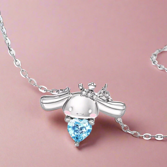 A silver necklace with a gold-plated chain, featuring a Cinnamoroll character pendant with a blue cubic zirconia in the center, displayed on a beach-themed background.